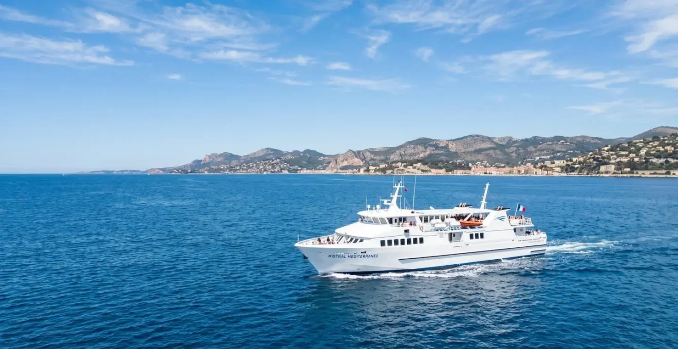 Un ferry blanc naviguant sur une mer Méditerranée calme et turquoise sous un ciel lumineux avec une côte visible au loin