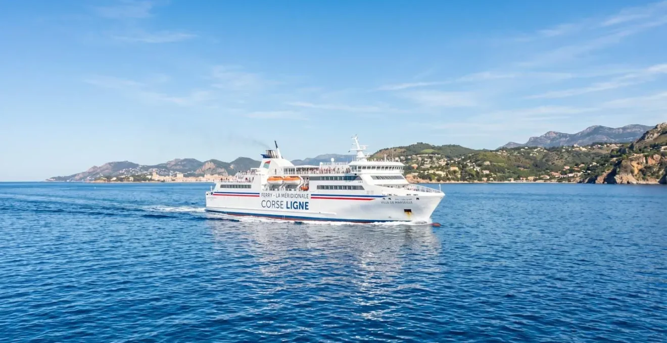 Un ferry blanc naviguant sur une mer Méditerranée calme et turquoise sous un ciel lumineux avec une côte visible au loin